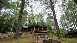 A cabin with a deck, surrounded by tall trees, and a fire pit with chairs in front. - Pinecone Place - A True North Vacation Home (Long Lake)