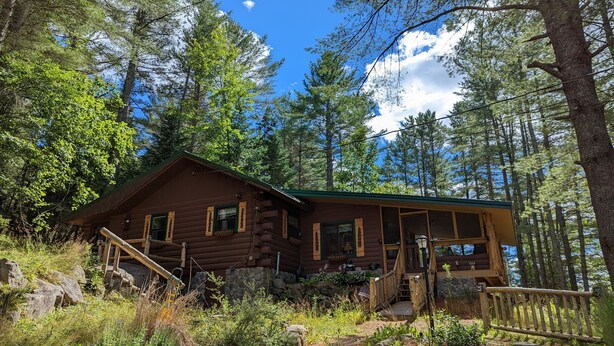 A rustic log cabin with a porch, surrounded by dense forest and a clear sky. - Pinecone Place - A True North Vacation Home (Long Lake)