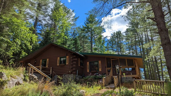 Un rustico chalet in legno con veranda, immerso in una fitta foresta sotto un cielo sereno.