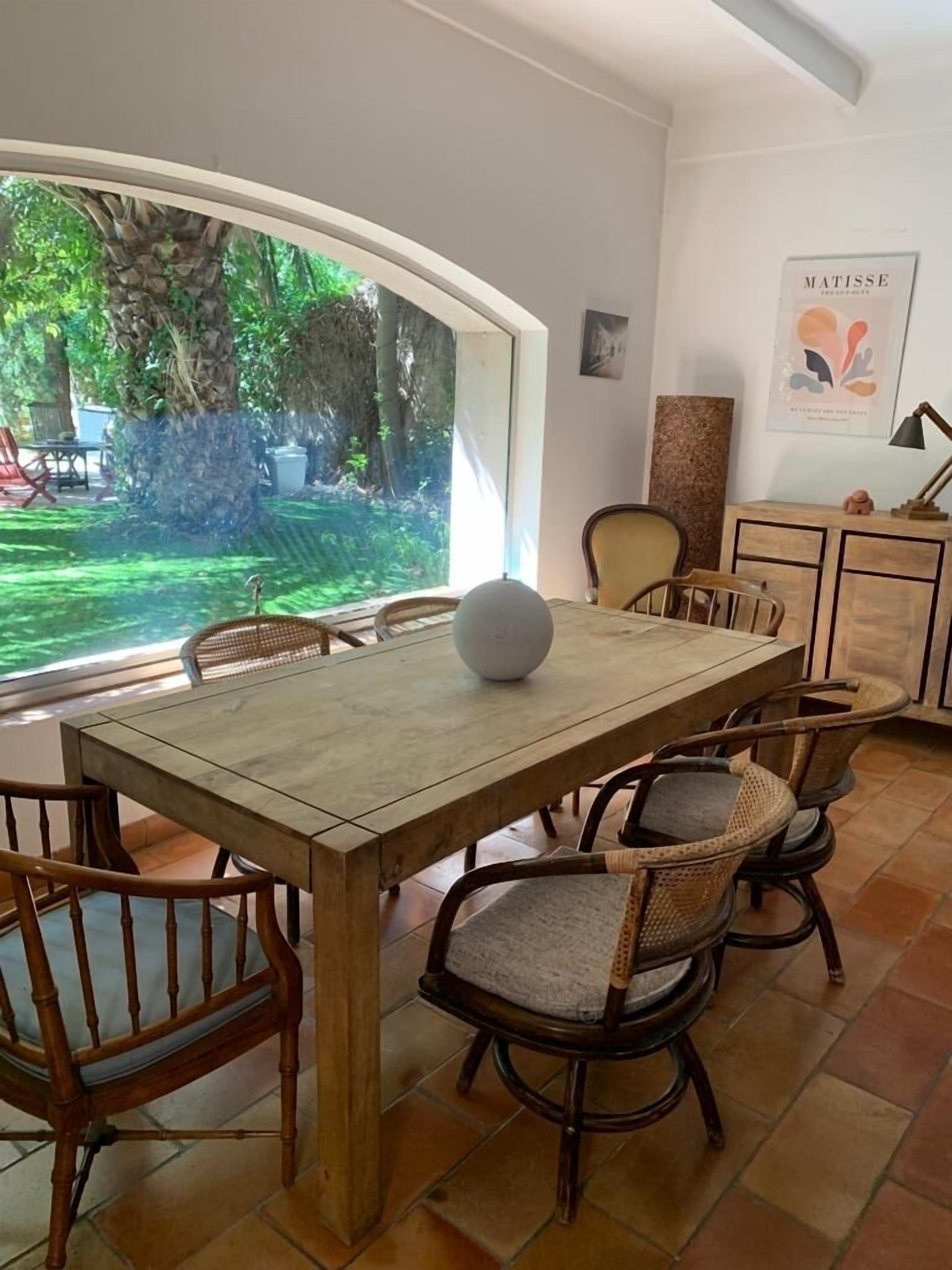 Un comedor con mesa de madera y sillas de mimbre, una esfera decorativa blanca sobre la mesa, y vistas a un jardín exuberante a través de amplios ventanales.