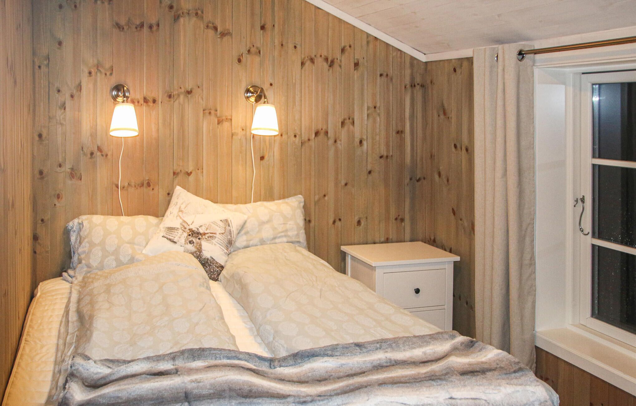 Een slaapkamer met een houten wand, een bed met beddengoed met een patroon, twee wandlampen, een nachtkastje en een raam met gordijnen.
