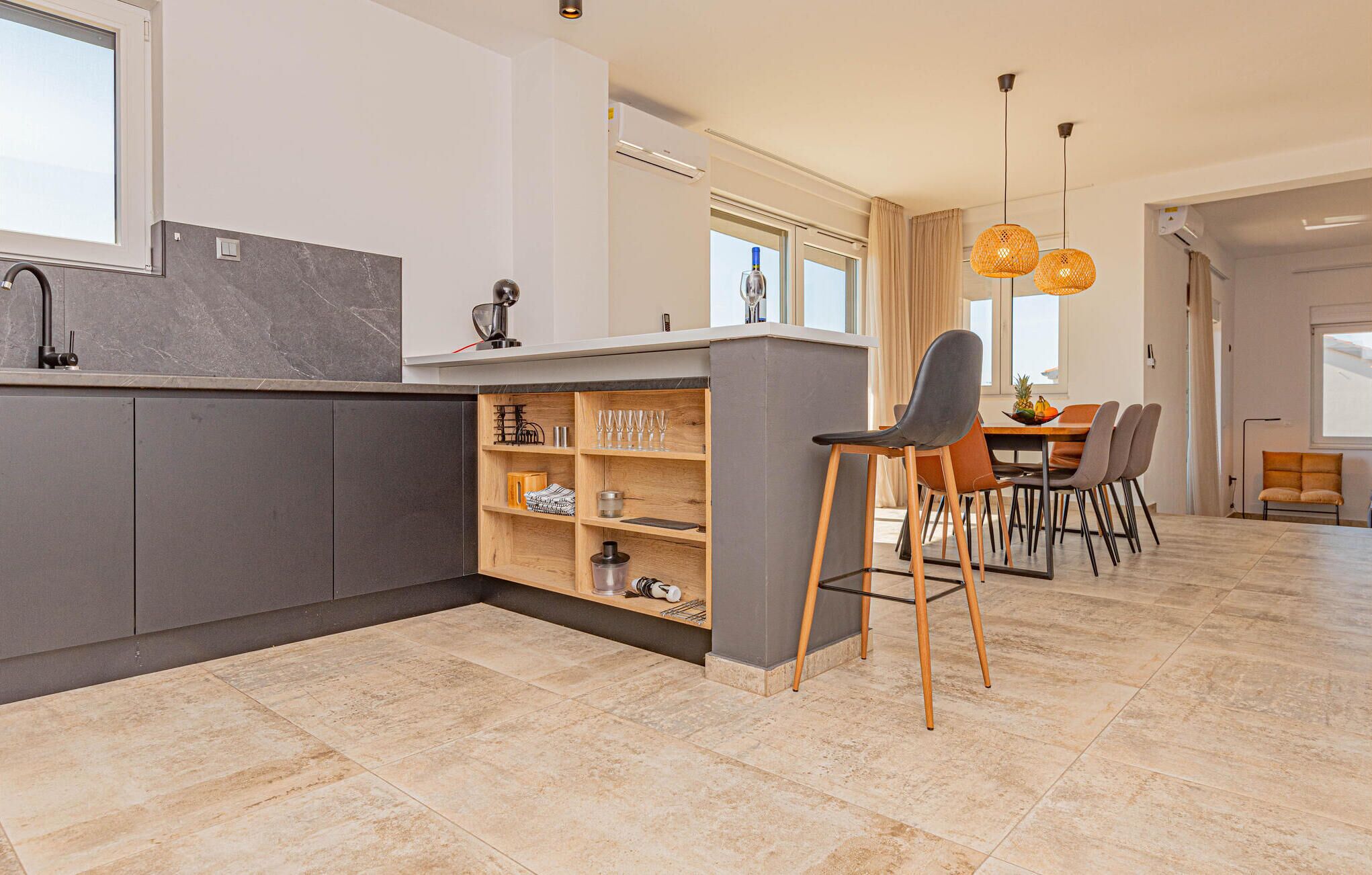 A modern kitchen with grey cabinets, a marble countertop, and a built-in wine rack. There is a dining area with a table and chairs, and a large window providing natural light.