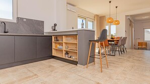 Una cocina moderna con armarios grises, encimera de mármol y una estantería empotrada para vinos. Hay una zona de comedor con mesa y sillas, y un ventanal que proporciona luz natural.