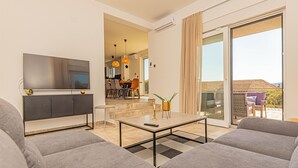 Una sala de estar moderna con un sofá, una mesa de centro y un televisor. Hay una puerta corrediza de vidrio que da a un balcón con mobiliario exterior.