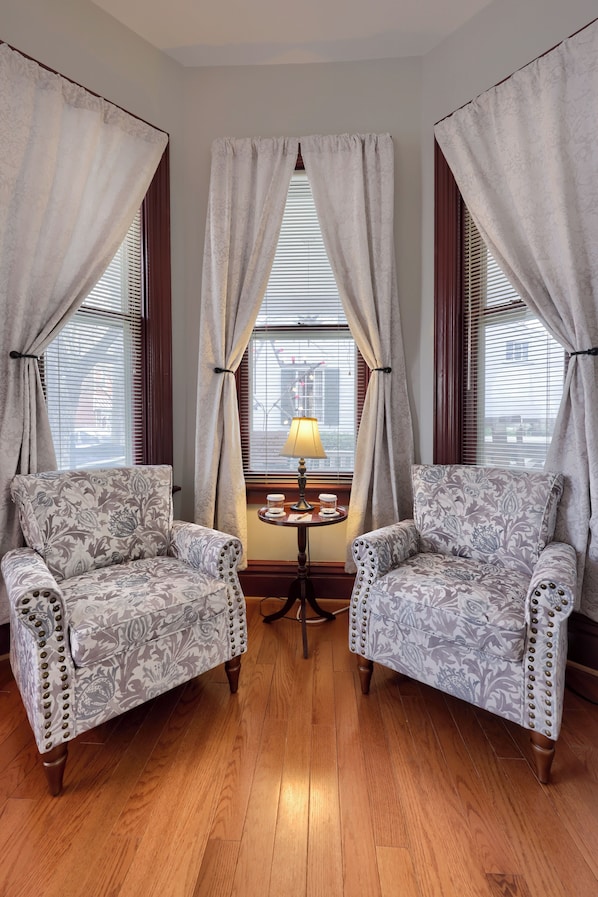 Two patterned armchairs facing each other with a small table between them, a lamp on the table, and a window with blinds in the background. - The Alden House (Lititz)