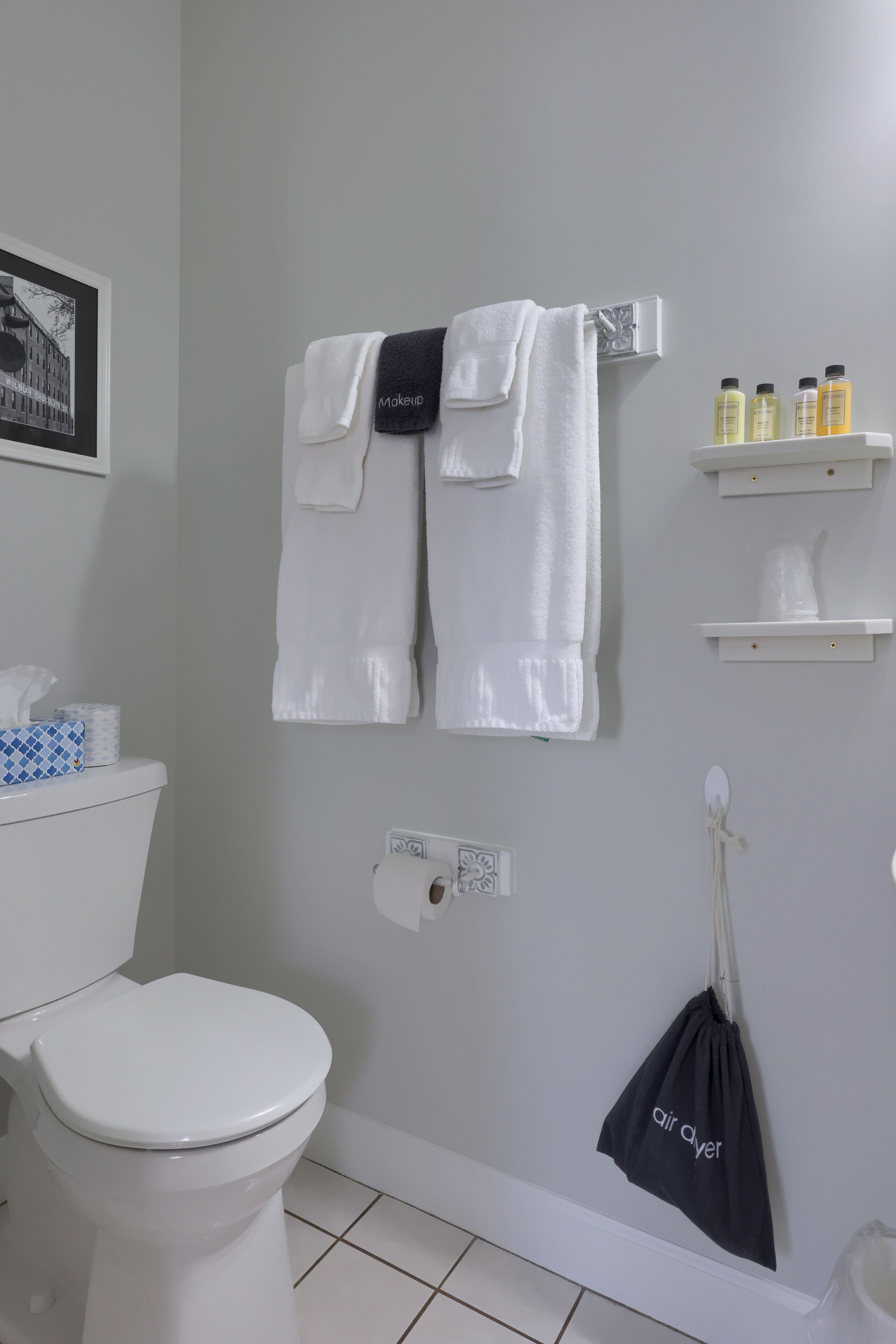 A bathroom with a toilet, two white towels hanging on a rack, a black bag labeled "Air Ayer," and a shelf with toiletries.