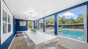 En overdækket terrasse med et hvidt bord og stole, store vinduer med udsigt over en swimmingpool og have.