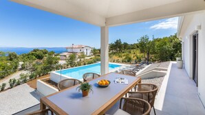 A balcony with a pool, wicker chairs, a table with a bowl of fruit, and a view of a house and landscape. - Amazing home in Labin (Labin)
