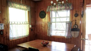 A wooden cabin interior with a dining table, two windows with curtains, a chandelier, and a stove. - Secluded Hillside Property with Valley Views and Fishing Pond (Burlington Flats)