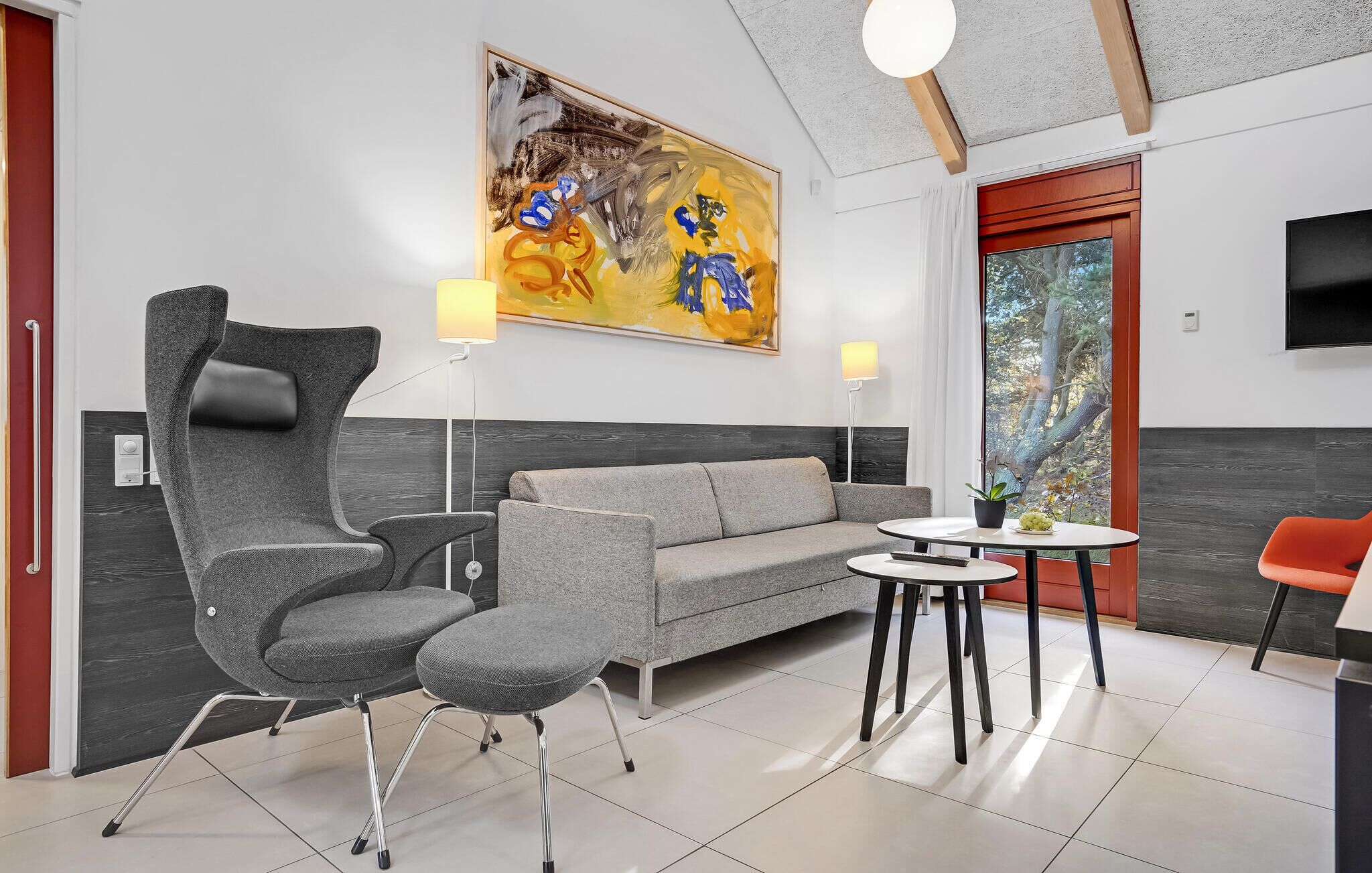 A modern living room with a grey sofa, two grey armchairs, a round coffee table, and a red accent chair. A large abstract painting hangs on the wall, and a window offers a view of trees.