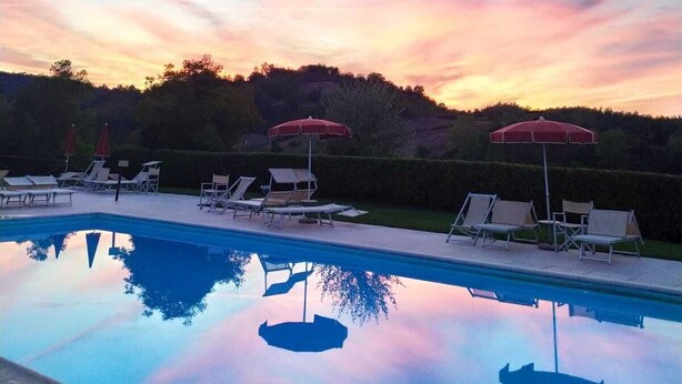 Une piscine entourée de chaises longues et de parasols, bordée d’une haie et d’arbres, le tout baigné par la lumière d’un ciel au coucher du soleil.