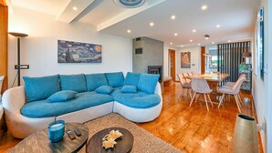 Un salon moderne comprenant un canapé d’angle, une cheminée, un coin repas avec une table et des chaises, et une grande fenêtre laissant entrer la lumière naturelle.