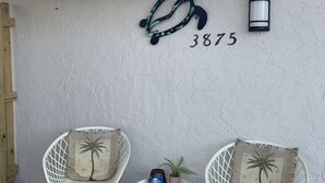 Pared exterior blanca con el número '3875' y un adorno metálico. Hay dos sillas blancas con cojines de hojas de palmera y una mesita con una planta y una botella azul.