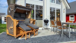 A wicker lounge chair set up outside a house with a large window, a barbecue grill, and a dining table with chairs. - Awesome home in Zerpenschleuse with sauna (Zerpenschleuse)
