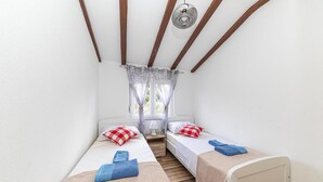 Una habitación pequeña con dos camas individuales, cada una con una almohada y una toalla. Hay una ventana con cortinas, una lámpara en una mesita de noche y vigas de madera en el techo.