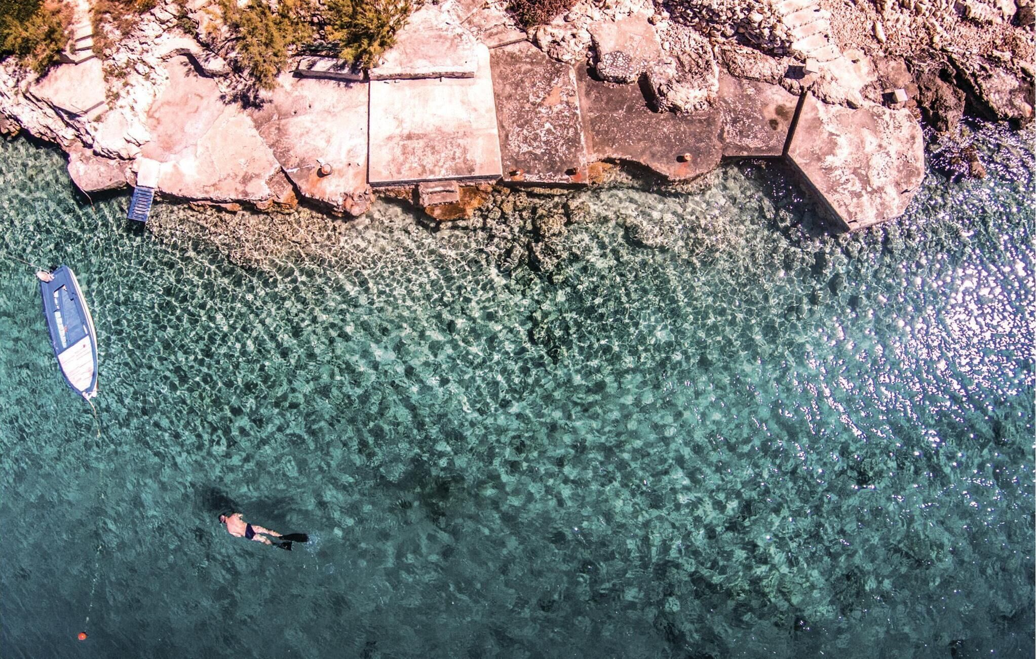 A clear view of a rocky shoreline with a boat anchored near the shore and a person swimming in the water.