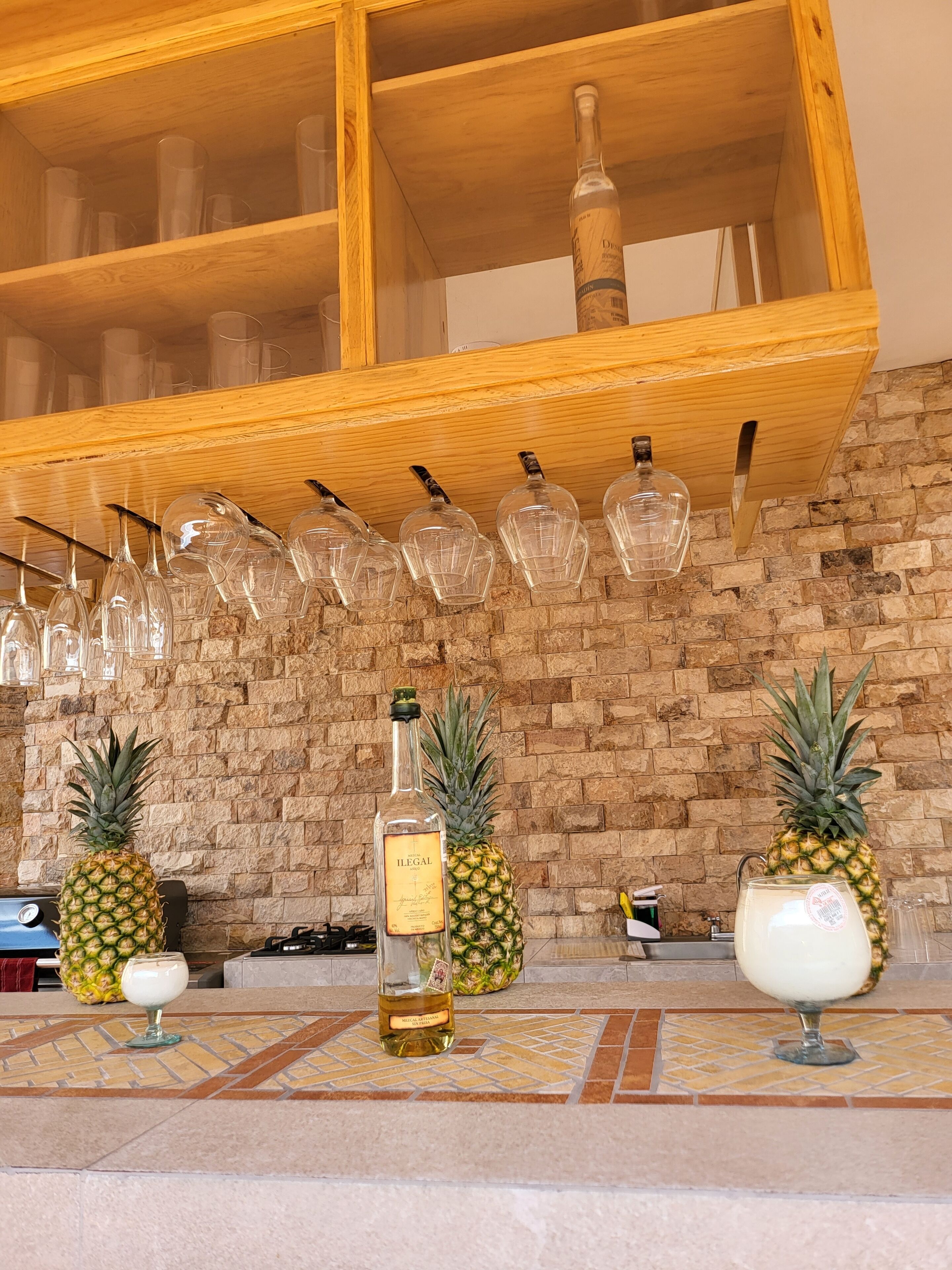 A bar area with a wooden shelf holding glasses, a bottle of liquor, and two pineapars on the counter.