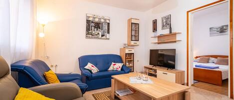 Een moderne woonkamer met een bank, fauteuil, salontafel, televisie en een bed op de achtergrond.