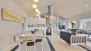 Un interior moderno con comedor, cocina y sala de estar, que presenta un techo revestido de madera, una chimenea central y un ventanal grande.