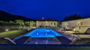 Ein rechteckiger Pool mit einem Steinhaus im Hintergrund, umgeben von einem Metallzaun und Bäumen.