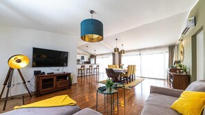 Ein modernes Wohnzimmer mit großem Flachbildfernseher, einem Essbereich mit Tisch und Stühlen sowie einer Küche im Hintergrund.