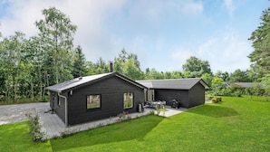 Ett modernt hus i två våningar med en mörk fasad, en uteplats med utemöbler och en välskött gräsmatta.