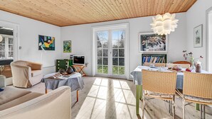 Un salon chaleureux avec un plafond en bois, un coin repas doté de meubles en osier et une bibliothèque remplie de livres.