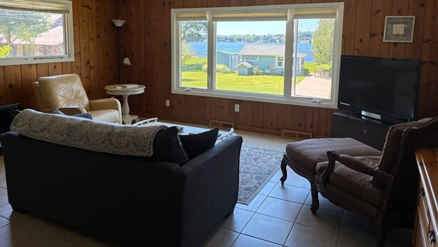 Sala de estar com ventoinha no teto, televisão, poltrona de couro, sofá e vista para uma casa e para a água lá fora.