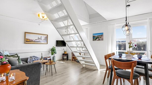 A modern living room with a staircase, a dining area with wooden chairs, a TV, and a large window with a view.