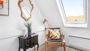 A room with a skylight, a wooden chair with a colorful cushion, a black side table with a plant, a gold-framed mirror, and a desk lamp.
