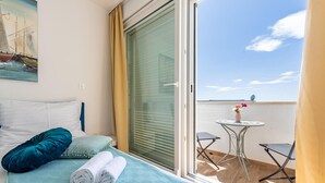 Une chambre à coucher avec un lit, une fenêtre avec des stores, un balcon avec une table et des chaises, et un tableau accroché au mur.