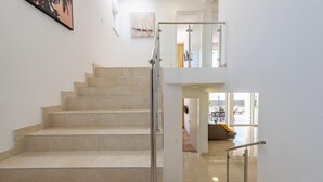 Un intérieur moderne avec un escalier menant à un deuxième étage, un coin repas avec des sièges extérieurs visibles à travers des portes vitrées, et des œuvres d'art accrochées aux murs.