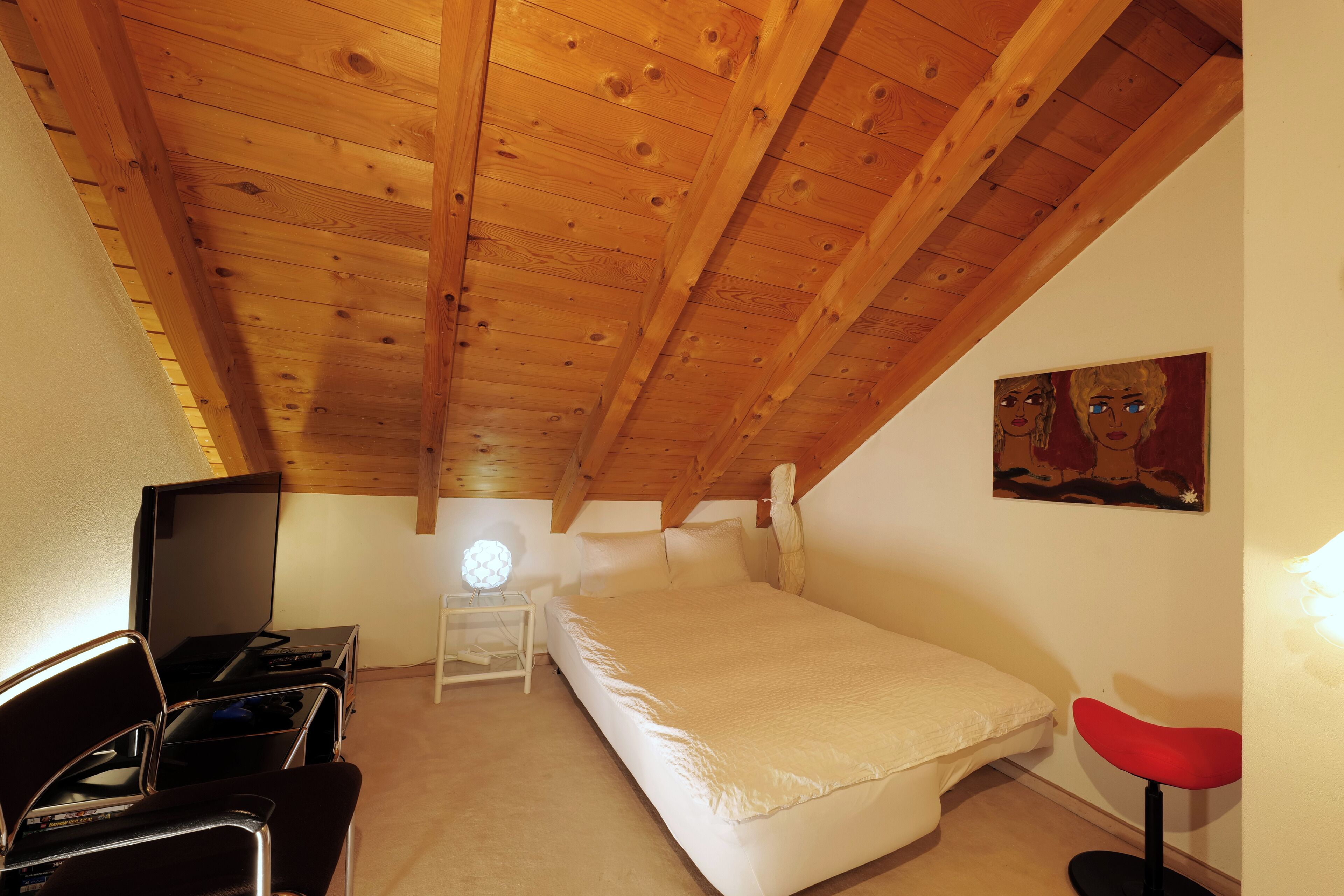 Una camera da letto con soffitto in legno, un letto, una scrivania con sedia, una TV e un quadro appeso al muro.