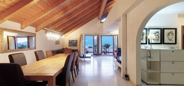 Uma ampla sala de jantar com uma mesa e cadeiras de madeira, um candelabro e vista para um pátio externo através de grandes portas de vidro.