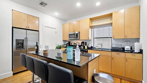 A modern kitchen with wooden cabinets, a stainless steel refrigerator, a microwave, and a built-in island with bar stools. - Houston Getaway by Downtown | Pool Table | Pets (Houston)