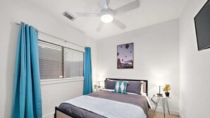 A bedroom with a bed, a ceiling fan, a window with curtains, a bedside table with a lamp, and a wall-mounted TV. - Houston Getaway by Downtown | Pool Table | Pets (Houston)