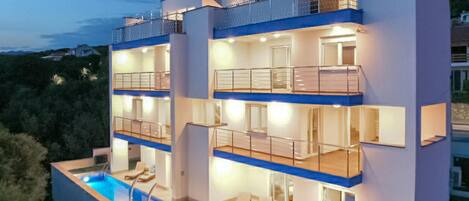 A multi-story modern building with balconies, a swimming pool, and a well-lit interior.