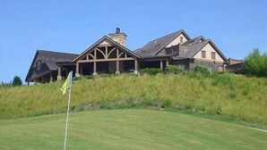 A large, two-story house with a stone facade and a wrap-around porch, situated on a grassy hill.
