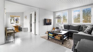A spacious living area with a grey sofa, a coffee table, and a TV mounted on the wall. There is a wooden desk with a chair, a small table with a lamp, and a window with a view of trees. - Beach front home in Nykøbing Sj with WiFi (Nykøbing Sj)