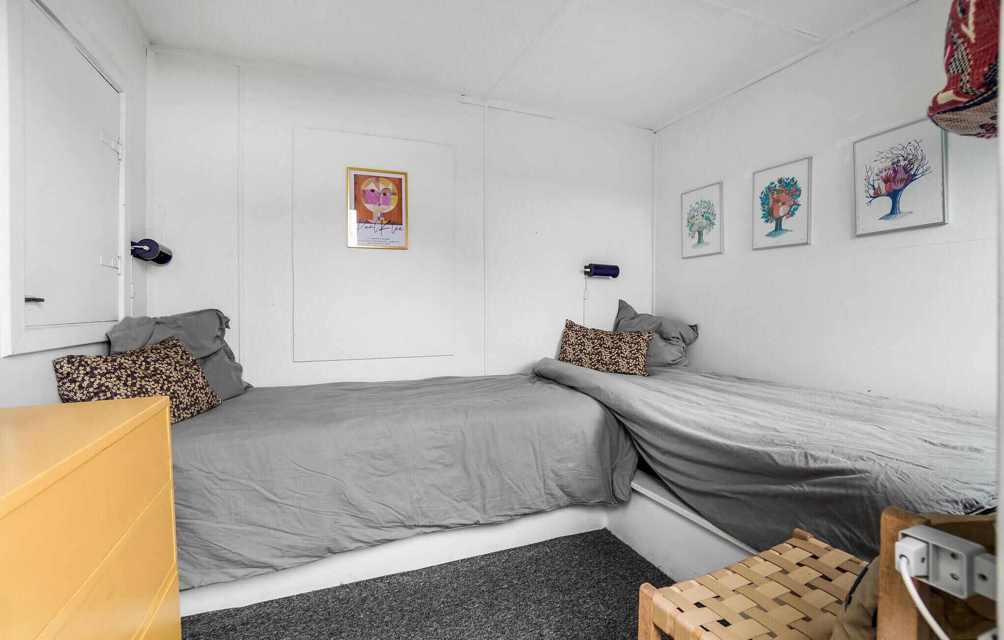 A bedroom with two single beds, a small wooden dresser, a desk, and a chair. There are framed pictures on the wall and a door with a poster.