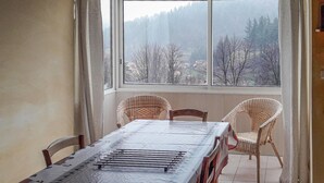 Ein Essbereich mit einem Tisch, der mit einer Schutzplane abgedeckt ist, zwei Korbstühlen und Blick auf einen Wald durch ein großes Fenster.