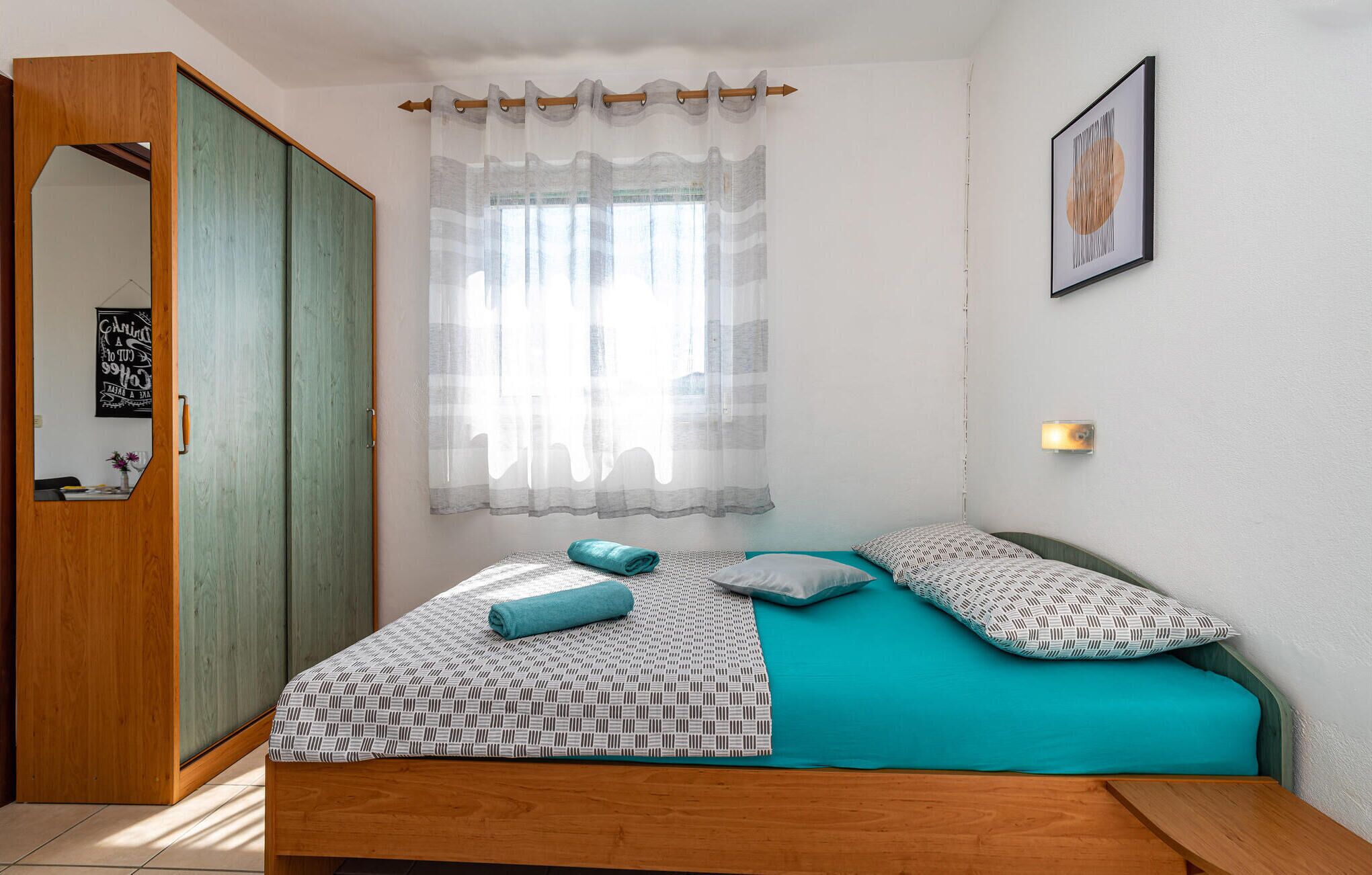 A bedroom with a bed, a wooden wardrobe, a window with sheer curtains, a wall-mounted light, and a framed picture.