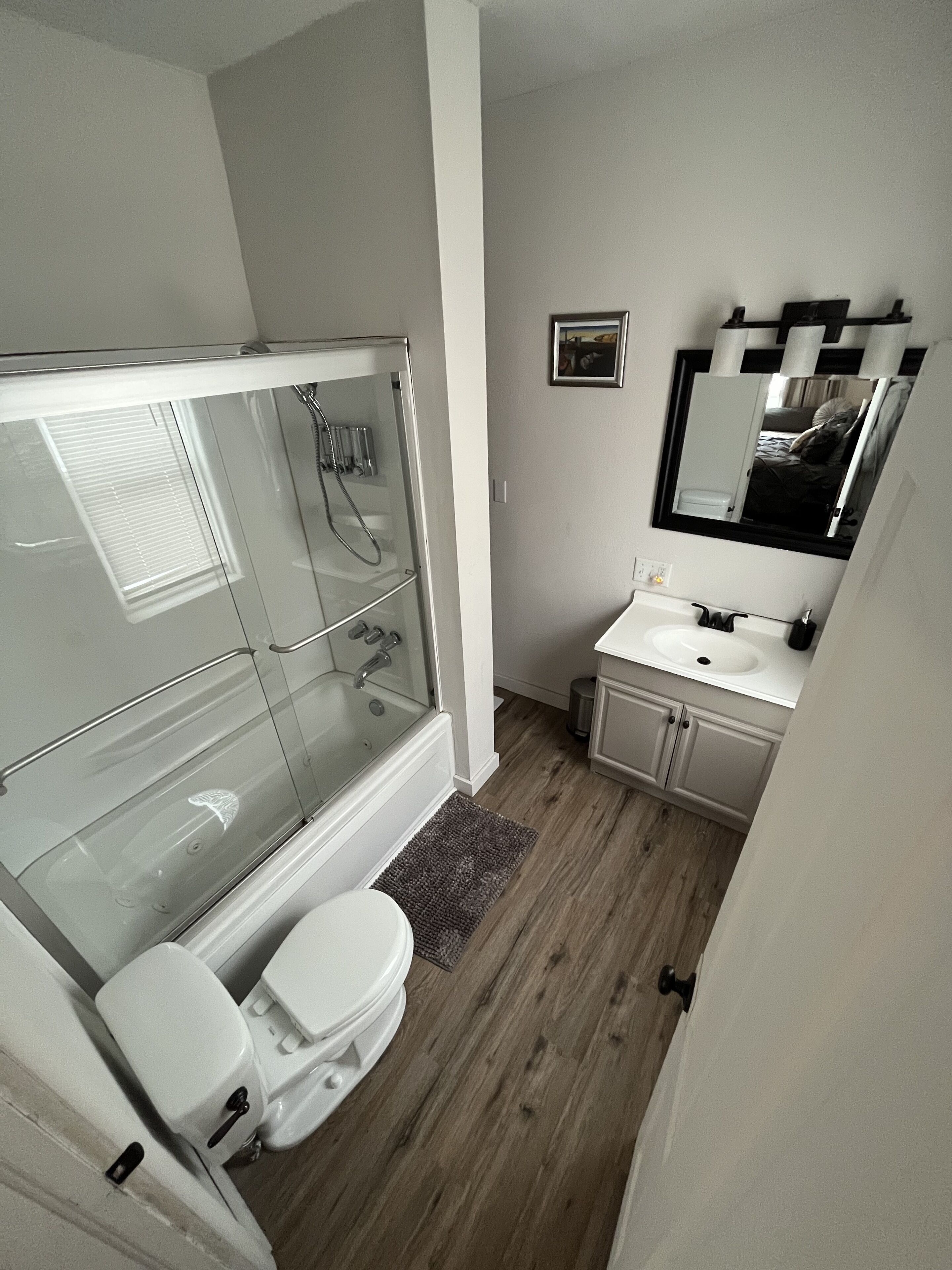 A bathroom with a glass-enclosed shower, a white toilet, a sink with a mirror above it, and wooden flooring.