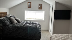 A bedroom with a sloped ceiling, a bed with a dark cover, a bedside table, a window with blinds, and a wall-mounted TV. - Spacious Historical Home in Downtown Philipsburg (Philipsburg)