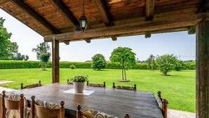 Une table en bois, entourée de chaises, placée sous une zone abritée, dans un jardin bien entretenu agrémenté de haies et d’arbres.