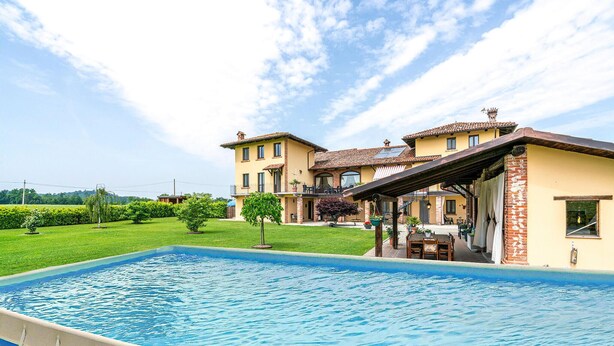 Une grande piscine, un patio couvert, une pelouse bien entretenue et un bâtiment à deux étages surmonté d’un toit en tuiles.