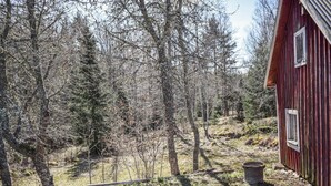 Une cabane rustique avec une porte et une fenêtre en bois, entourée d’une zone boisée avec des arbres et une colline herbeuse.