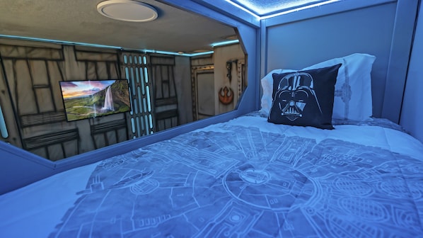 Une chambre avec un grand lit, un oreiller orné d’un motif de Dark Vador et un téléviseur fixé au mur qui diffuse un paysage pittoresque.
