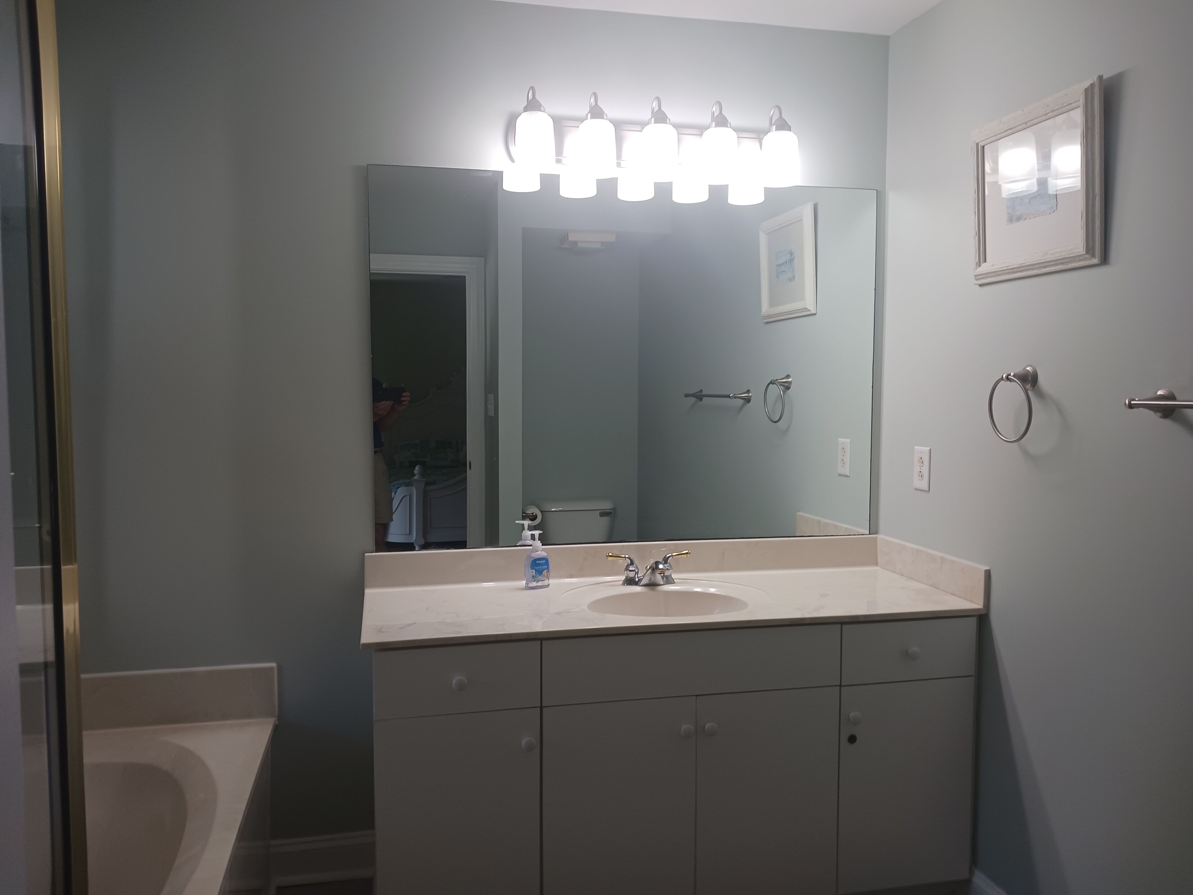 Ein Badezimmer mit einem großen Spiegel, einem Waschbecken mit Armatur und einer Toilette. An der Wand befinden sich Handtuchhalter, und über dem Spiegel hängt ein gerahmtes Bild.