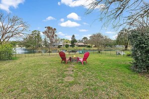 A fenced backyard with two red chairs and a fire pit, surrounded by trees and a lake view. - *Malibu Cottage on Lake Mandarin* WIFI, FIRE PIT (Jacksonville)